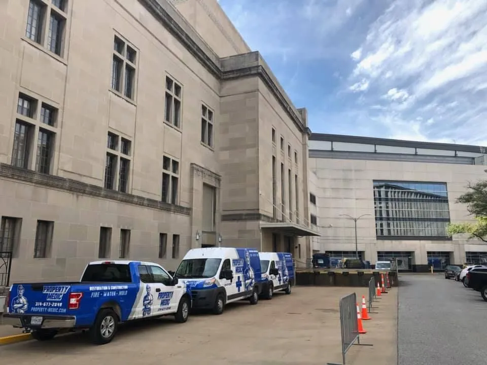 Three blue and white home repair and restoration service vehicles are parked outside a large, beige institutional building on a partly cloudy day. Orange traffic cones line the street.